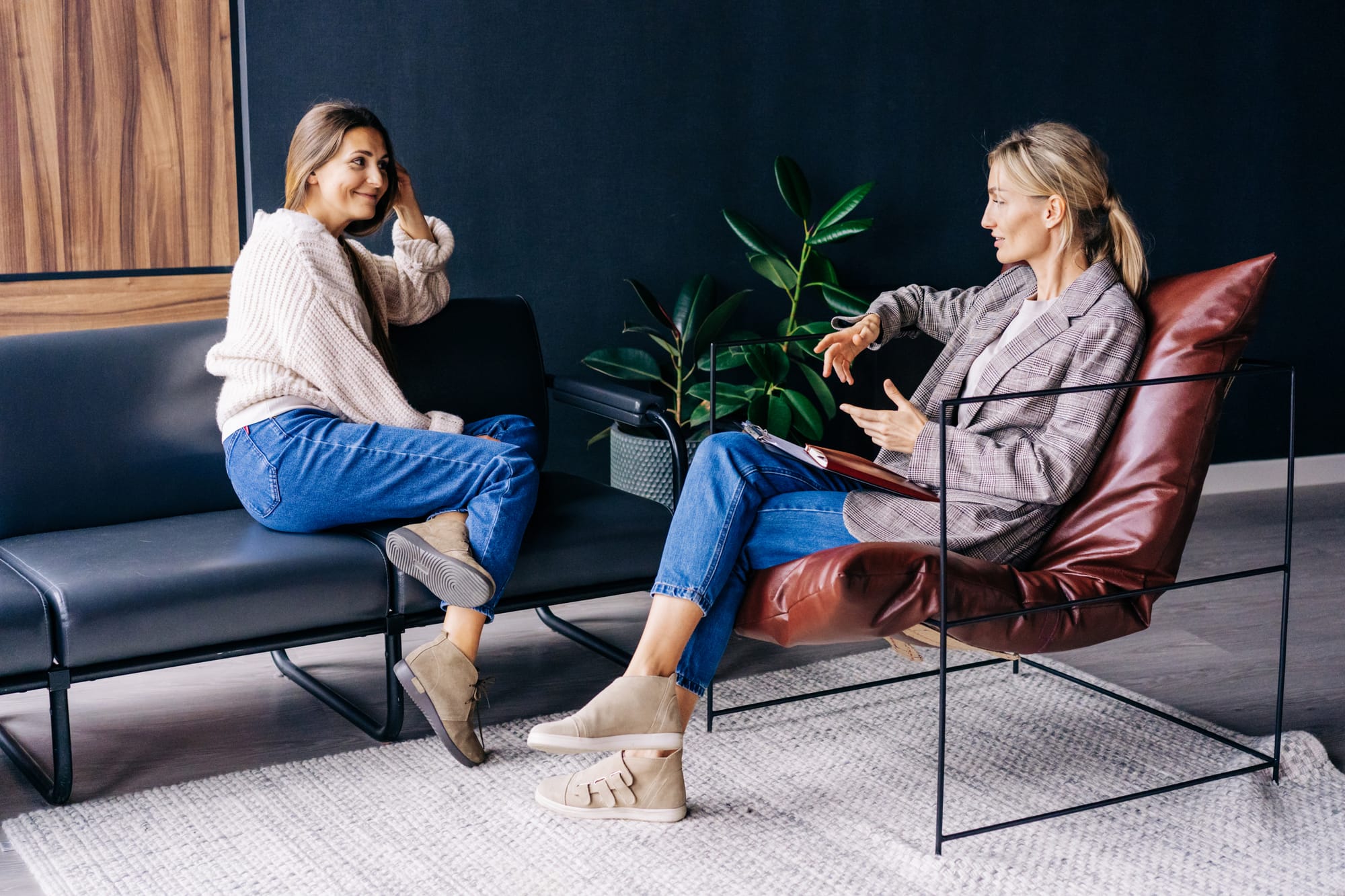Two women talking in a modern therapy office, one seated on a dark sofa and the other in a leather chair with a notebook.