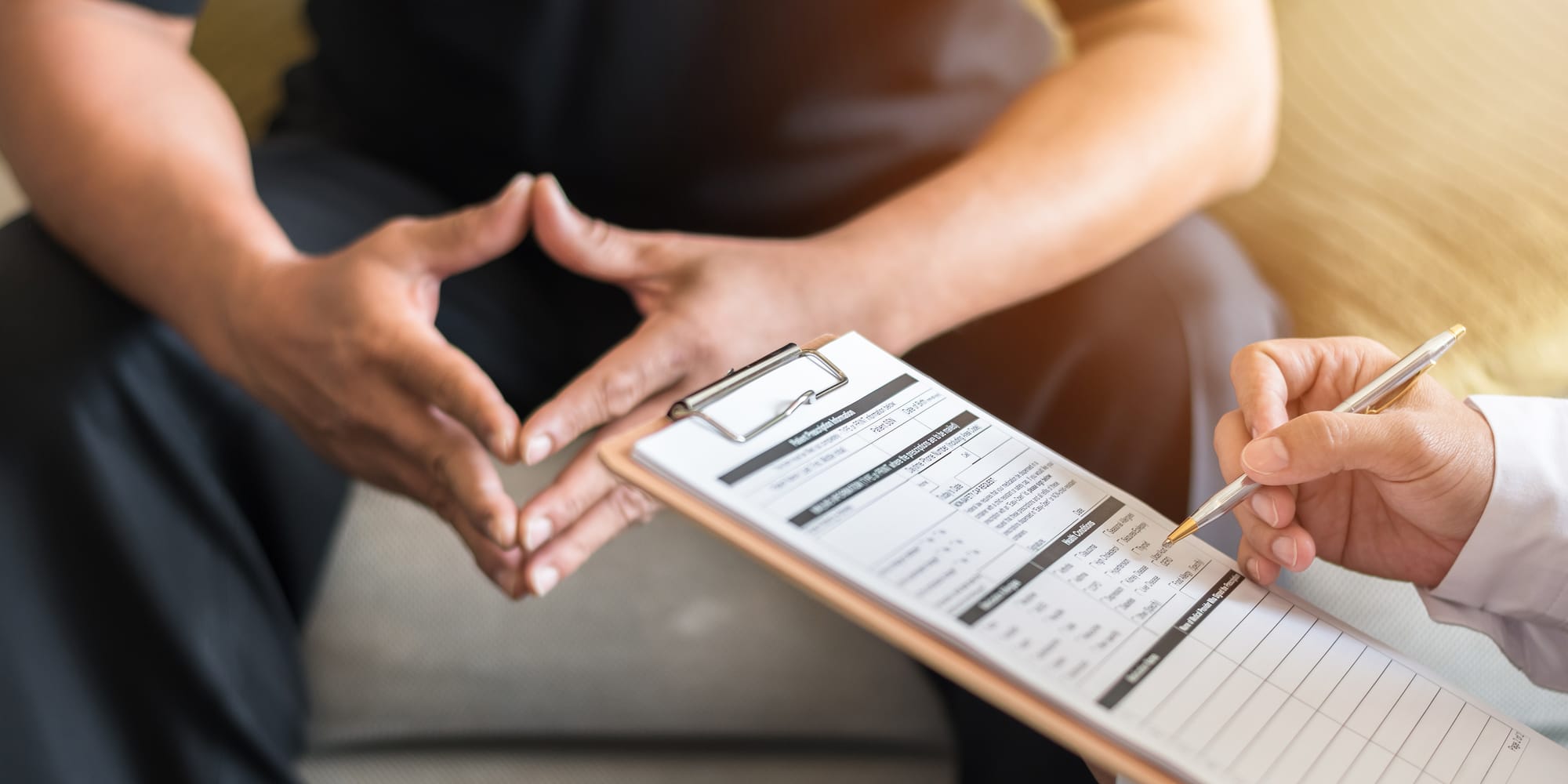 A clinician holds a clipboard with an insurance form while a patient gestures during a warm, softly lit conversation.
