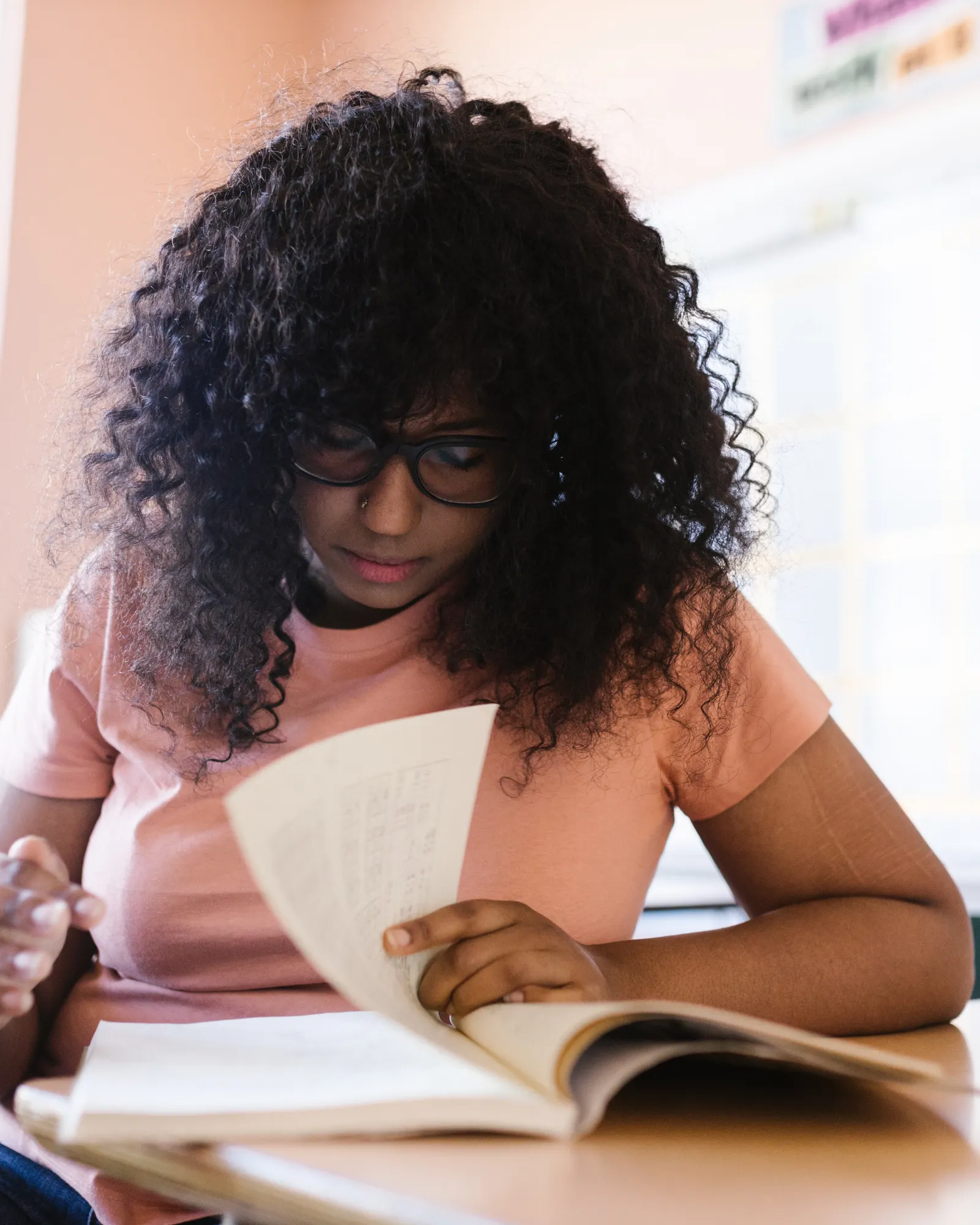 A high school student reading a book at her desk in a bright classroom, focused on the page.