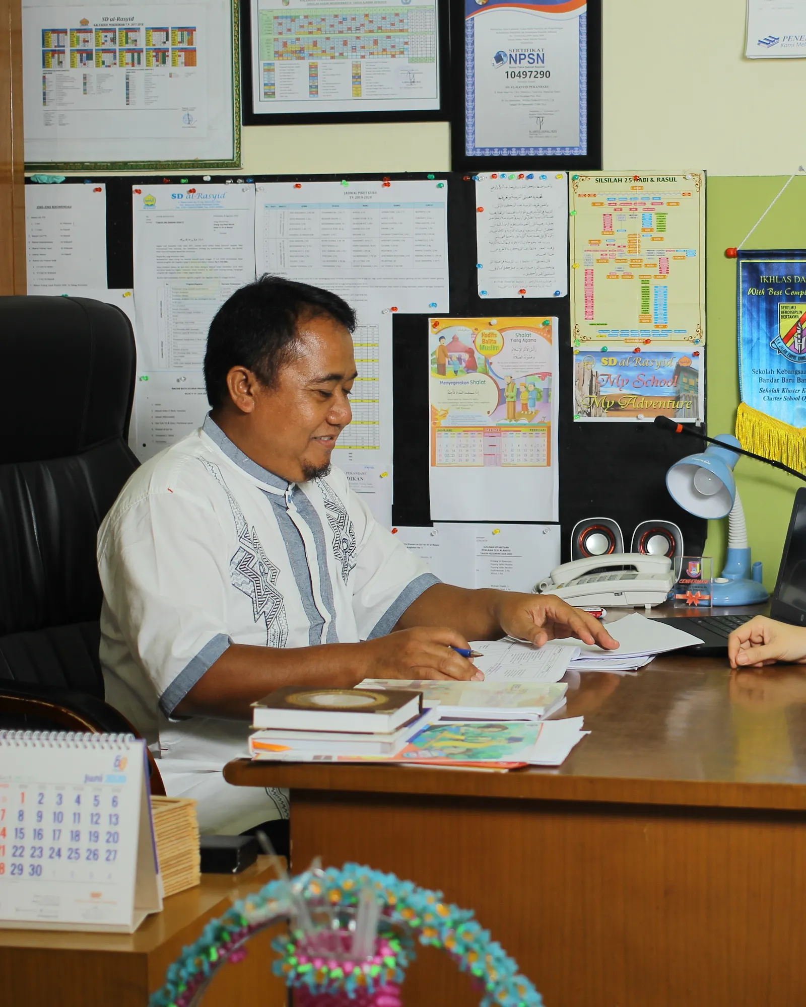 A teacher and a parent sitting together at a desk in a school office, talking over paperwork.