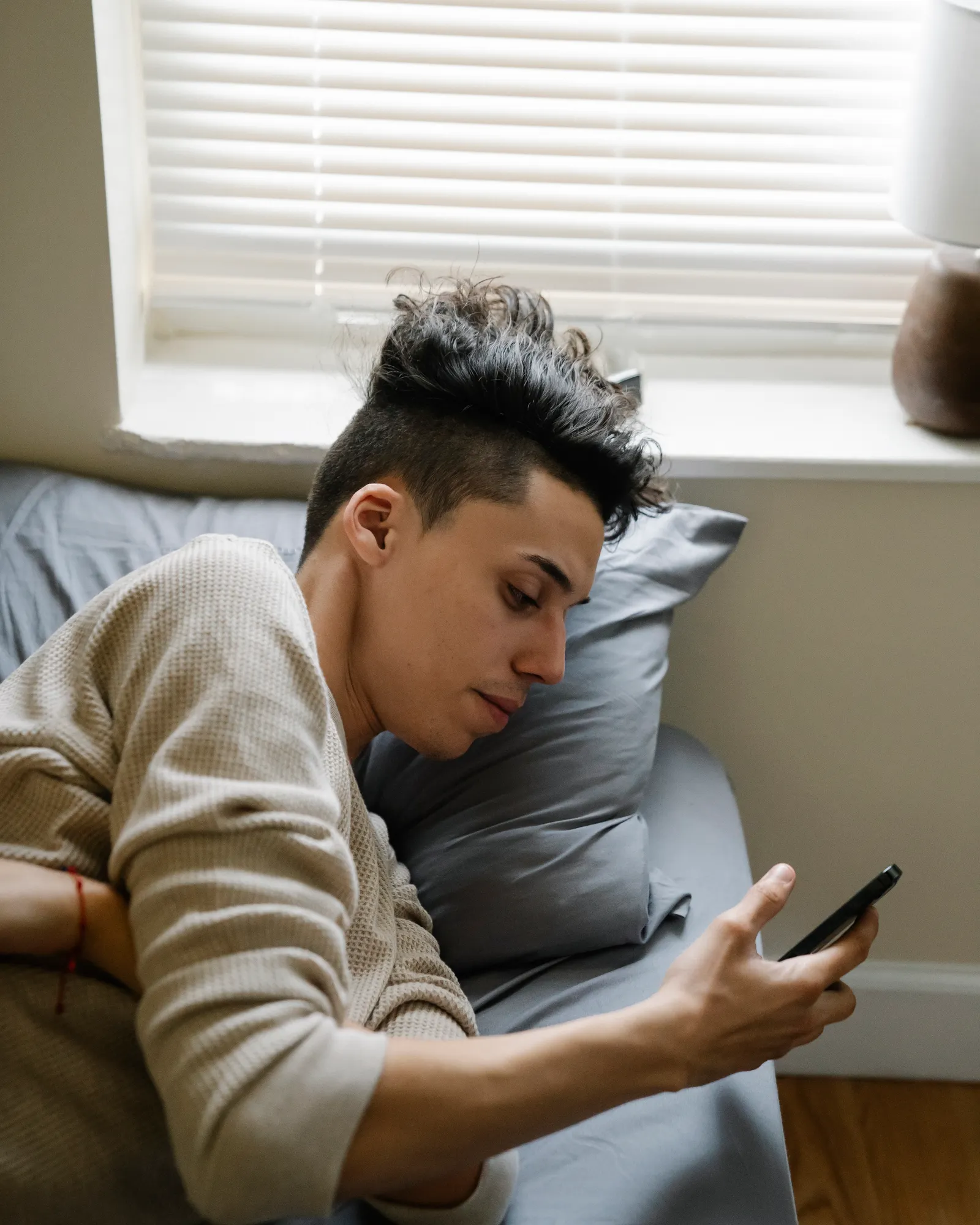 A teenage boy lying on his bed in morning light, scrolling on his phone.