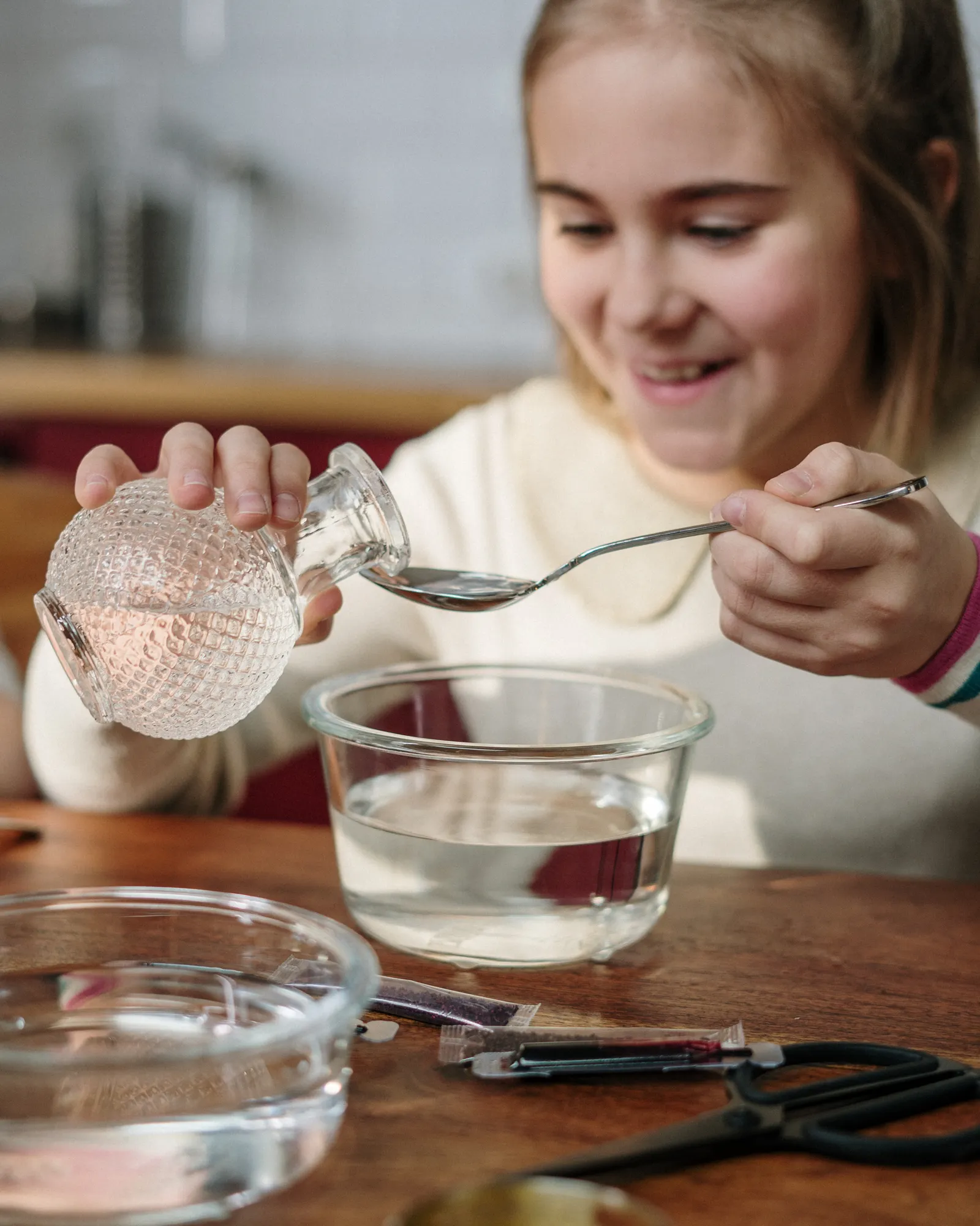 A young girl at a wooden kitchen table, absorbed in a hands-on water and color experiment.
