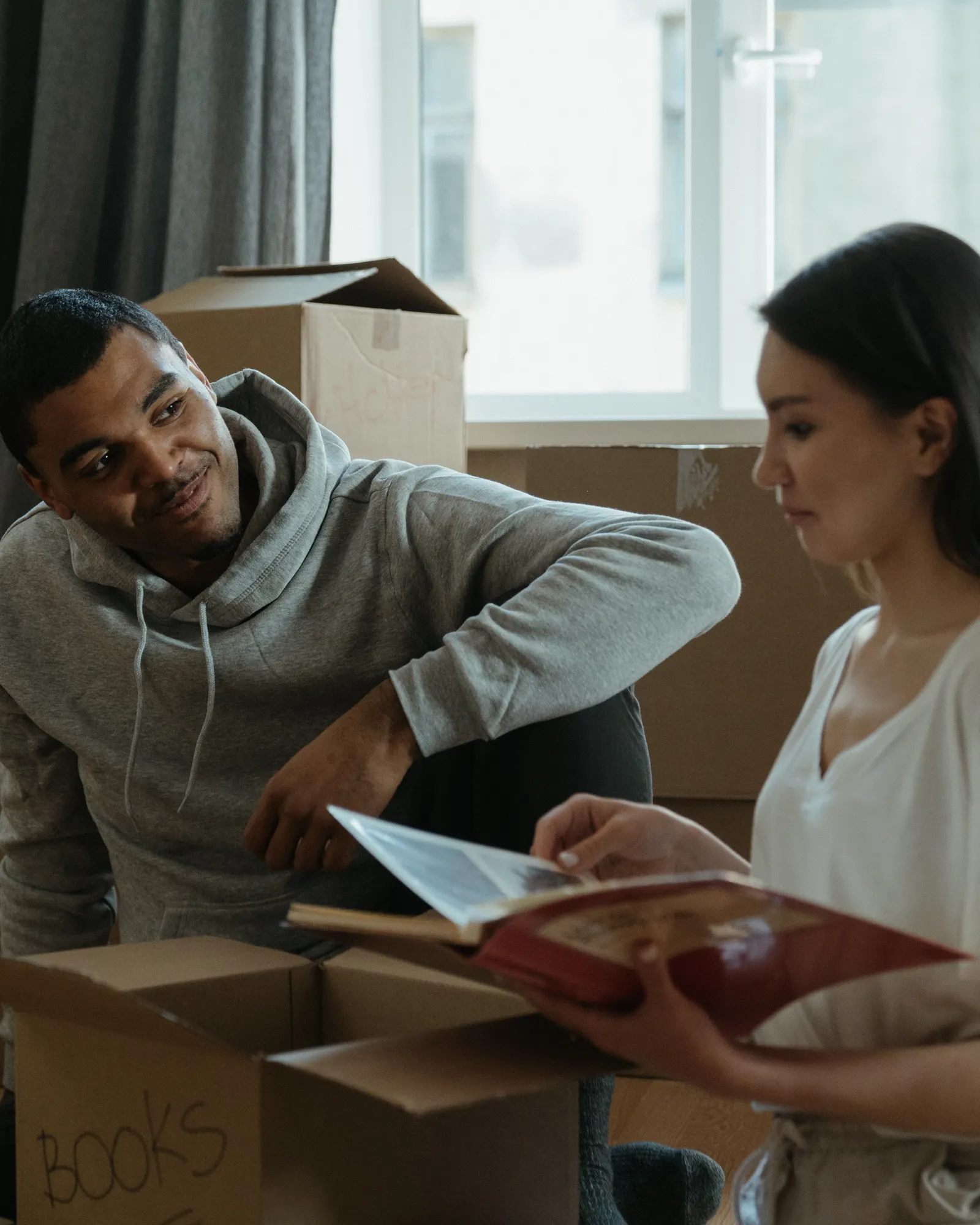 A couple in a bright room unpacking moving boxes and pausing over a photo album together.