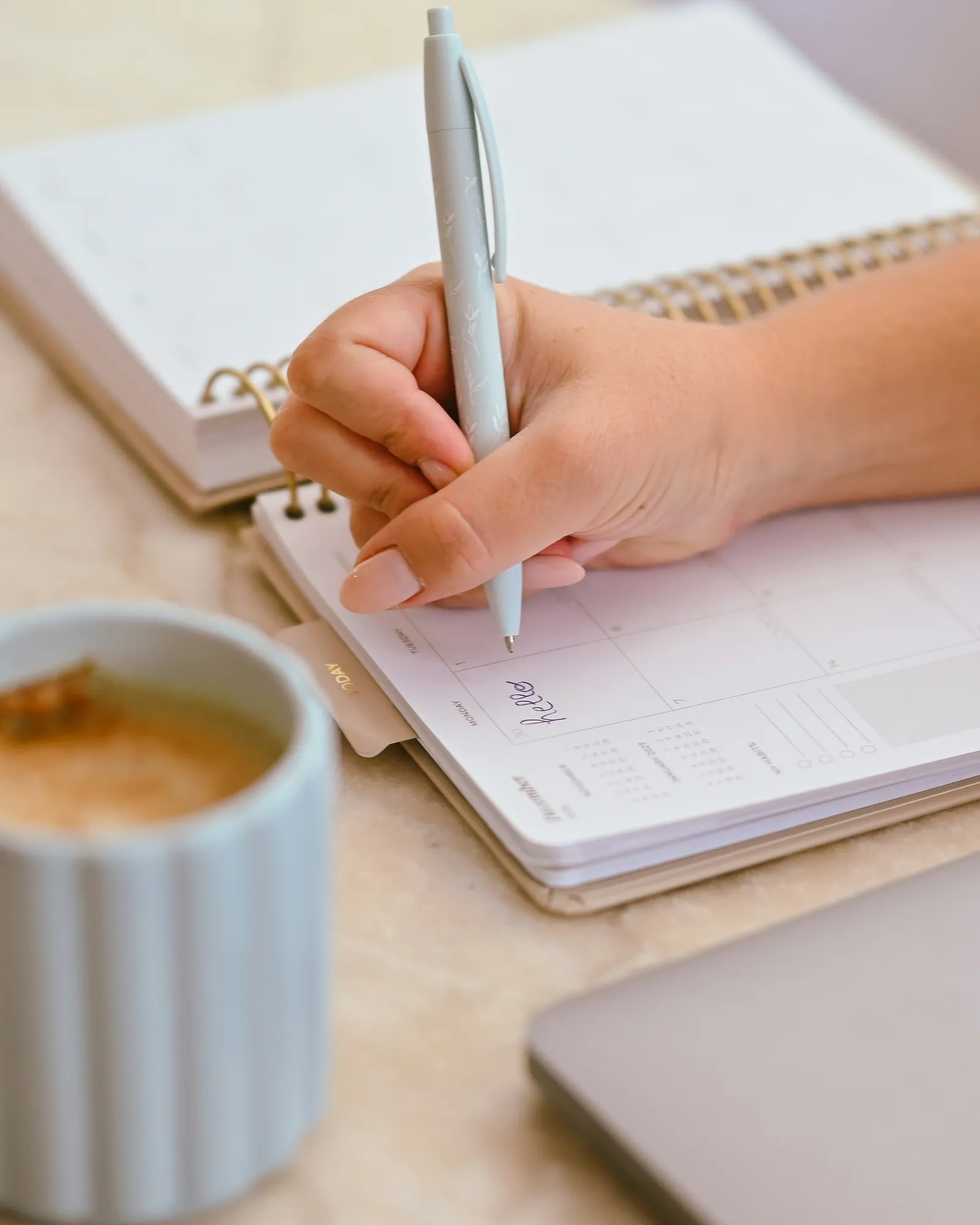 A person writes in a paper planner with a light blue pen next to a coffee mug.