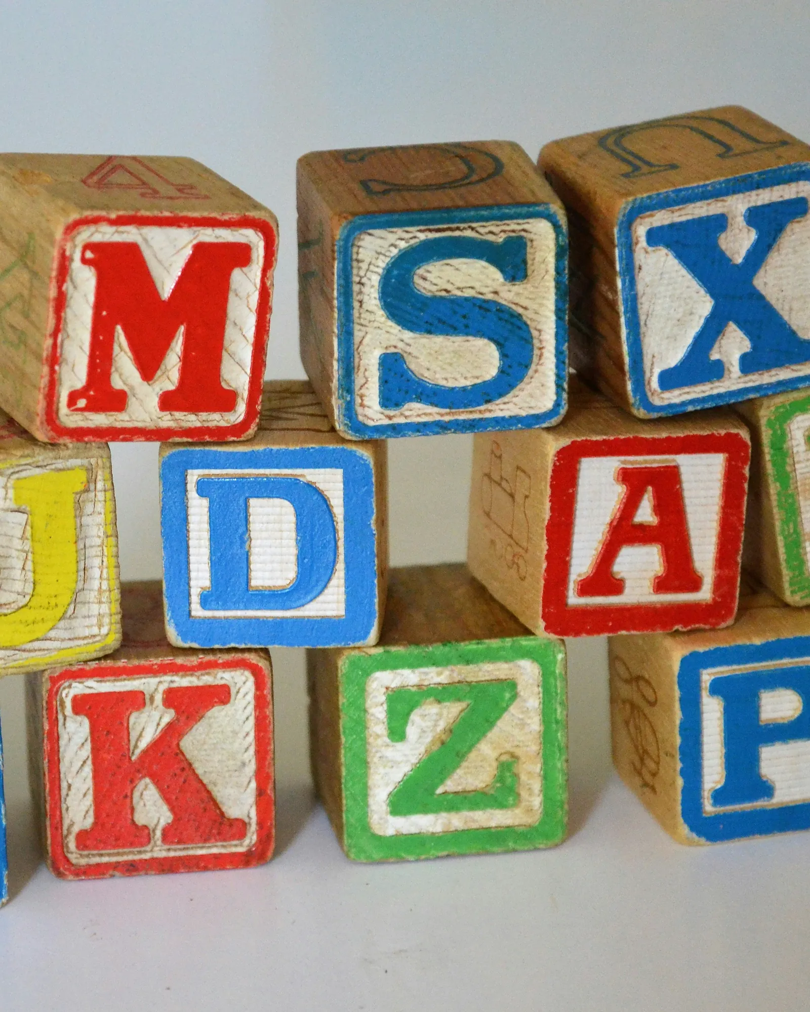 Wooden alphabet blocks with red, blue and green letters are stacked upright on a white surface.
