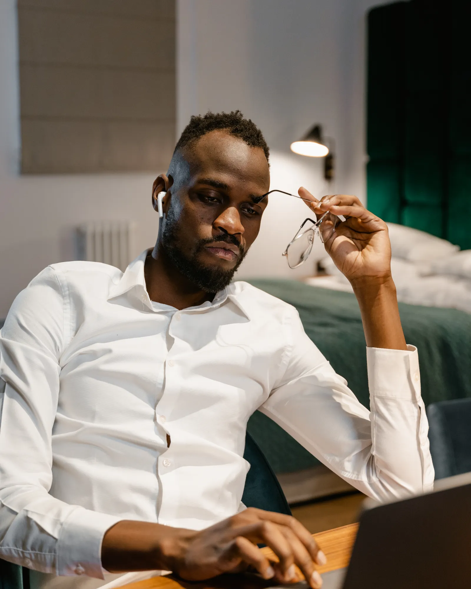 A man sits at a table at home with a laptop, holding his glasses as he reads the screen.