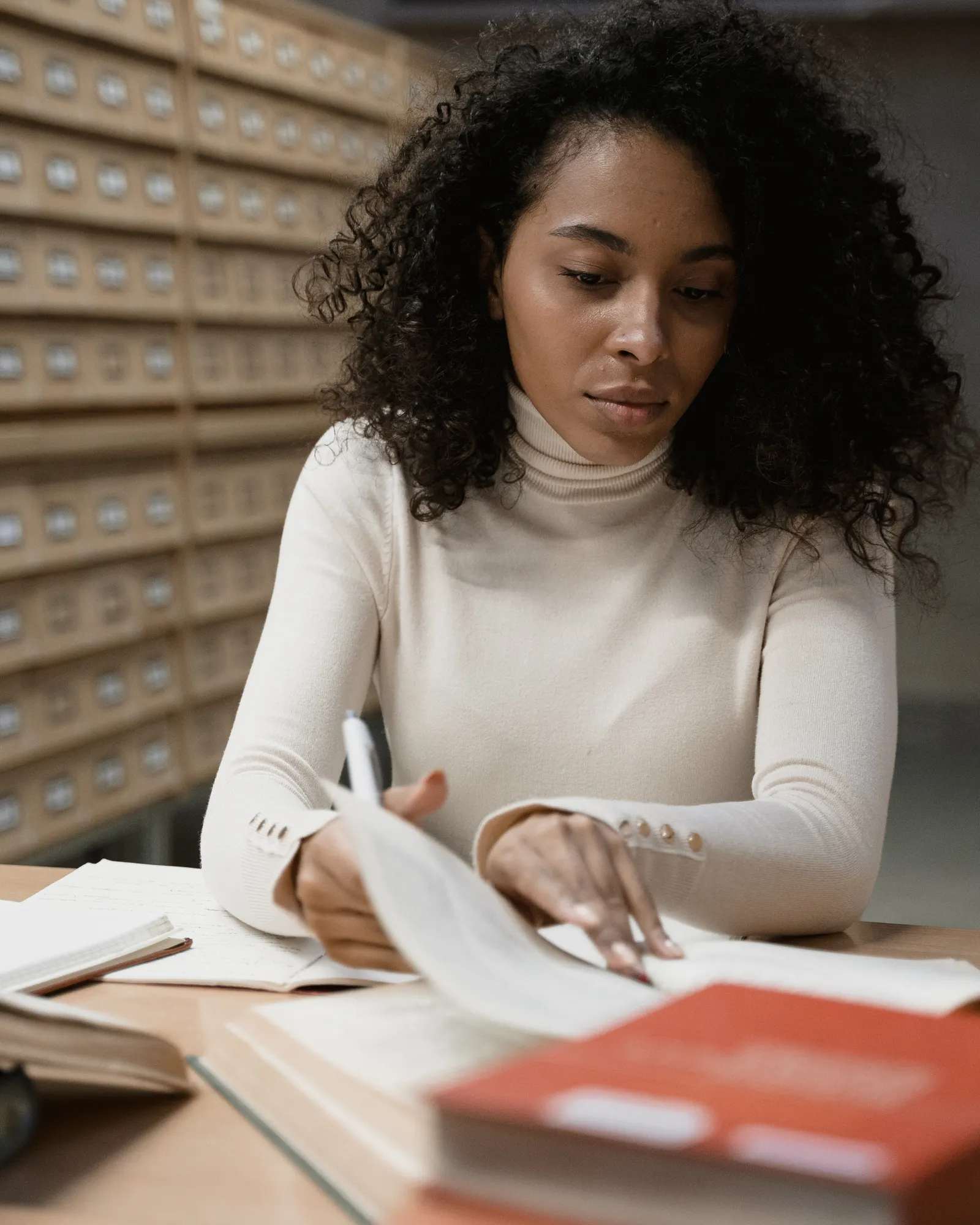 A young woman sits at a library desk with several open books in front of her.