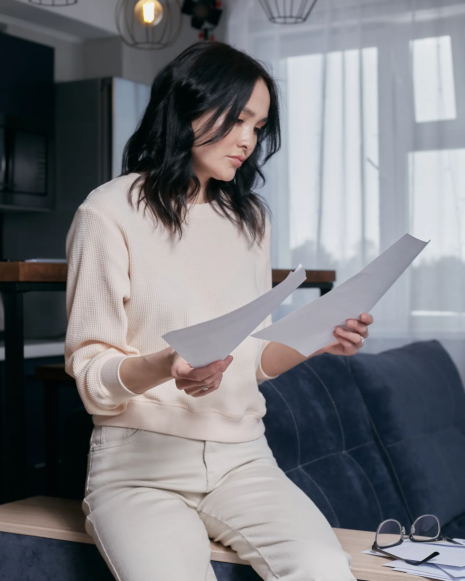 A woman in glasses sits at home reading through a printed multi-page document.