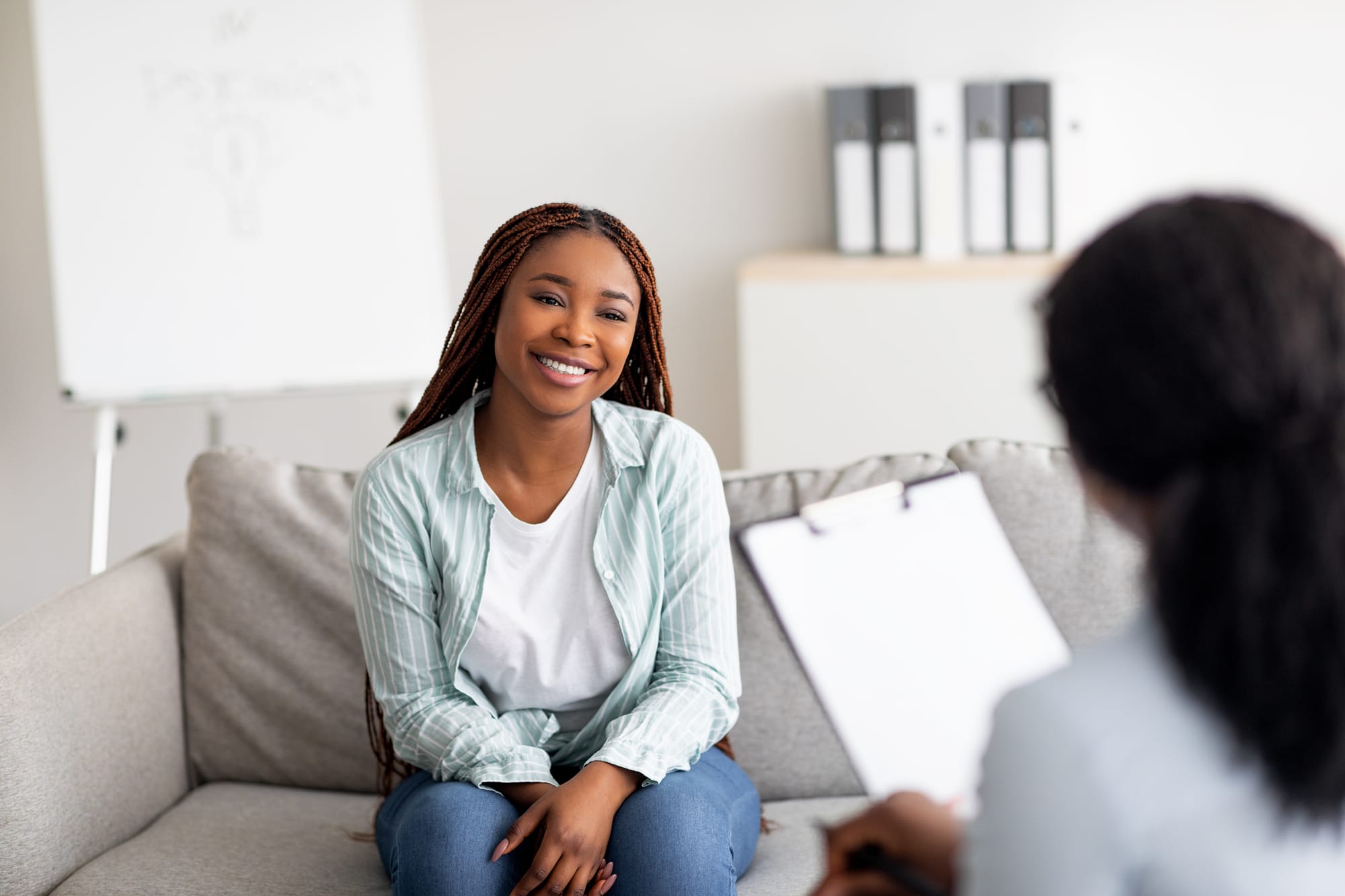 A smiling woman sitting on a couch in a bright therapy office, talking with a clinician holding a clipboard.